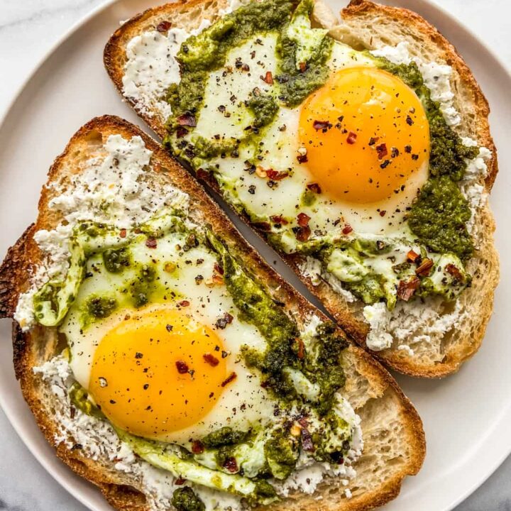 Sunny-side-up eggs cooked in pesto on 2 slices of bread on a white plate.